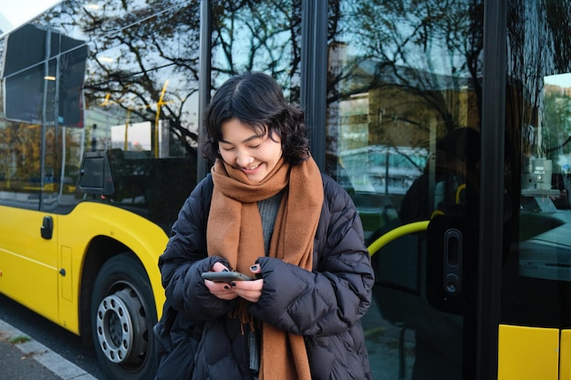 Free photo beautiful korean girl student on bus stop looking at her smartphone checking timetable reading text