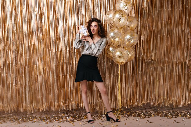 Beautiful lady in festive outfit holding gift on background of golden balloons