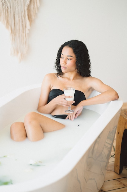 Beautiful lady thinking and holding a glass of milk while having a milk bath .