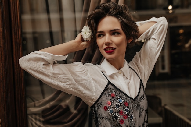 Beautiful lady with curly hair and red lips looking away inside. Cool brunette woman in light blouse holding flowers posing in cafe.