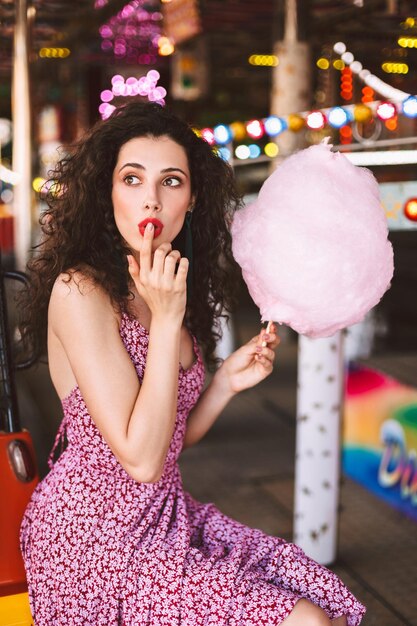 Beautiful lady with dark curly hair in dress sitting with cotton candy in hand and dreamily looking aside while spending time in amusement park