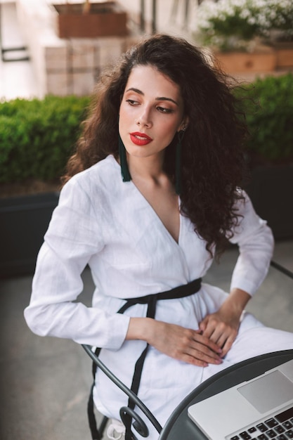 Beautiful lady with dark curly hair in white costume sitting at the table with  laptop and dreamily looking aside in cafe on street