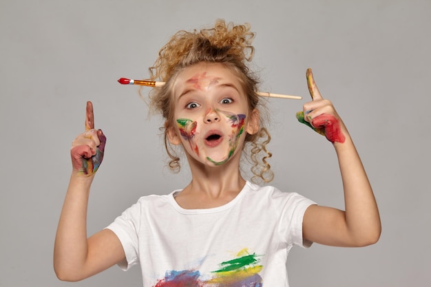 Free photo beautiful little girl having a brush in her chic curly blond hair, wearing in a white t-shirt, with a painted fingers, is acting like she has got an idea, on a gray background.