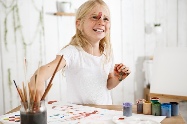 Free photo beautiful little girl with straight blonde hair, freckles and paint on her face, laughing and having fun. kids' art activities.