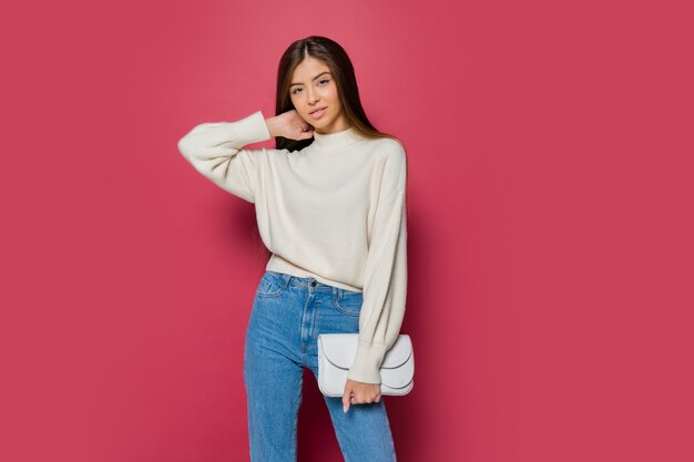 Beautiful long haired  woman in cozy white pullover  and casual jeans  posing on pink background isolate. Holding  eco leather hand bag.