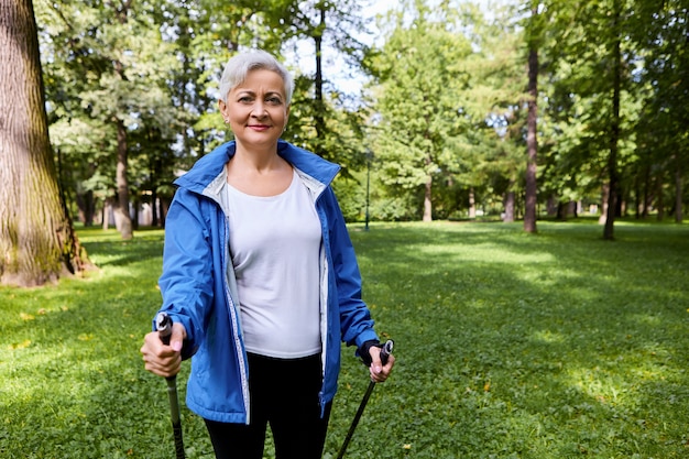 Free photo beautiful mature sportswoman enjoying her active hobby, holding sticks for scandinavian walking, having fit body, spending retirement days on healthy activity. summertime, sports and leisure