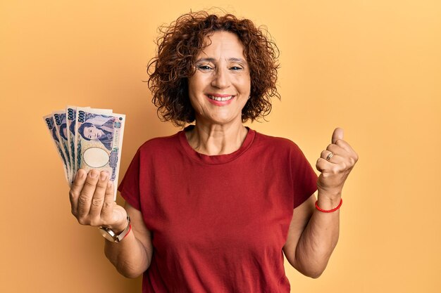 Beautiful middle age mature woman holding 5000 japanese yen banknotes screaming proud, celebrating victory and success very excited with raised arm