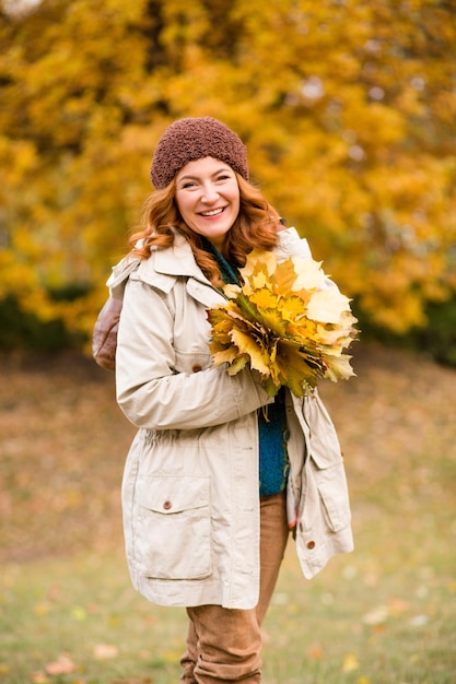 Beautiful middle-aged woman walking in autumn park. Smiling woman holding many yellow maple leaves and looking at the camera.