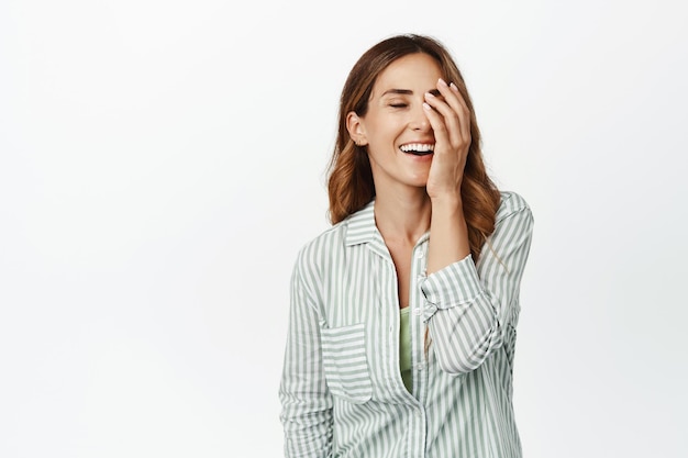 Beautiful middle aged woman with perfect skin, no wrinkles, smiling and laughing, touching face, standing in blouse against white background