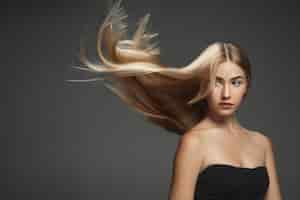 Free photo beautiful model with long smooth, flying blonde hair isolated on dark grey studio background. young caucasian model with well-kept skin and hair blowing on air.