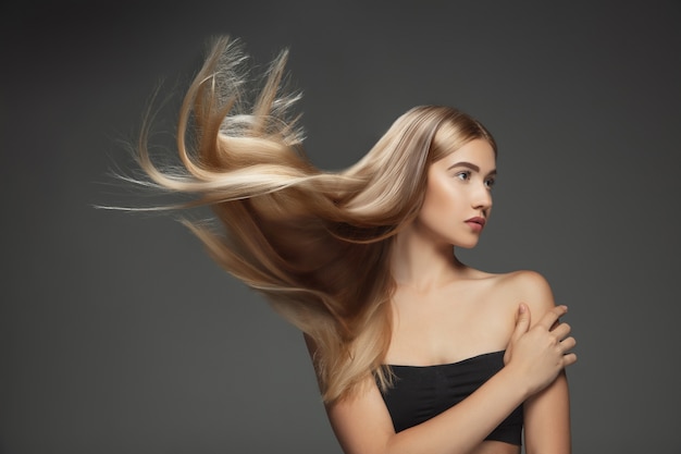 Beautiful model with long smooth, flying blonde hair isolated on dark grey studio background. Young caucasian model with well-kept skin and hair blowing on air.