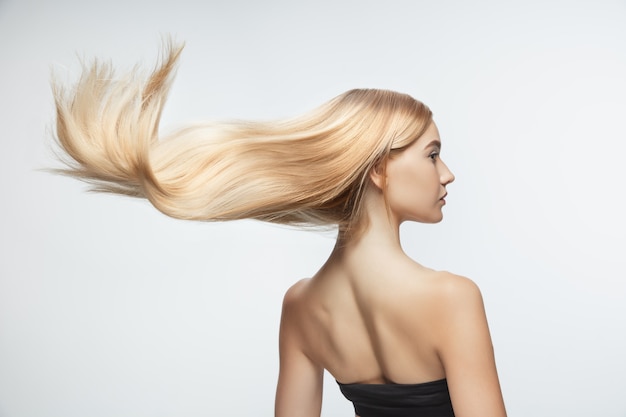 Free photo beautiful model with long smooth, flying blonde hair isolated on white studio background. young caucasian model with well-kept skin and hair blowing on air.