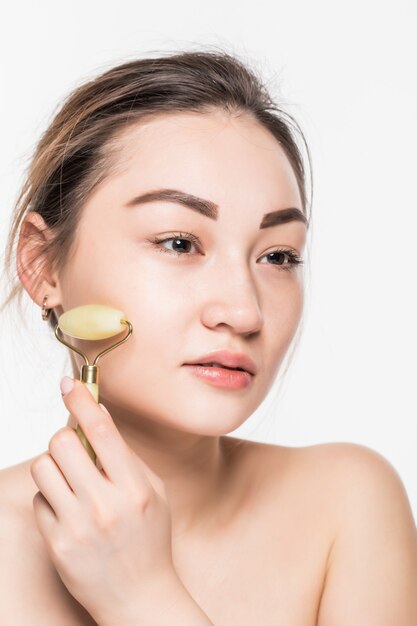 Beautiful model woman with healthy fresh clean skin enjoying a massage with a jade face roller to improve circulation, relax the muscles and tone the skin, isolated on gray wall with copy space