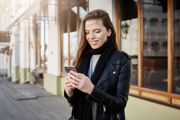 Beautiful modern woman in trendy clothes holding smartphone and looking at screen while messaging or browsing in net, walking on street