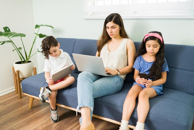 Beautiful mom typing on the laptop and working while sitting on the couch with her son and daughter. Little kids playing with a tablet and smartphone