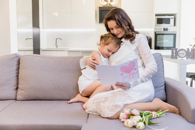 Beautiful mother and daughter sitting on sofa in living room reading greeting card