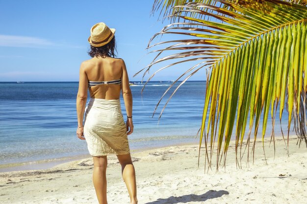 Beautiful picture of a brunette female in a swimsuit next to a palm against blue lagoon at daytime