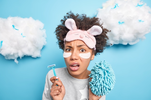 Beautiful puzzled woman holds razor and sponge going to have daily hygiene routines dressed in pajama and sleepmask applies beauty patces under eyes isolated on blue wall