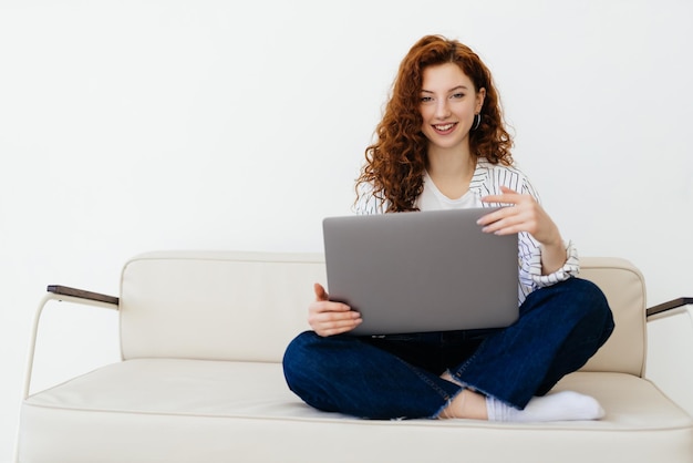 Beautiful redhaired woman working online using laptop sitting on comfortable grey couch