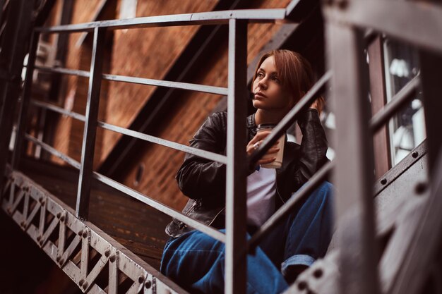 A beautiful redhead girl wearing trendy clothes sitting on stairs outside the cafe and holding takeaway coffee.