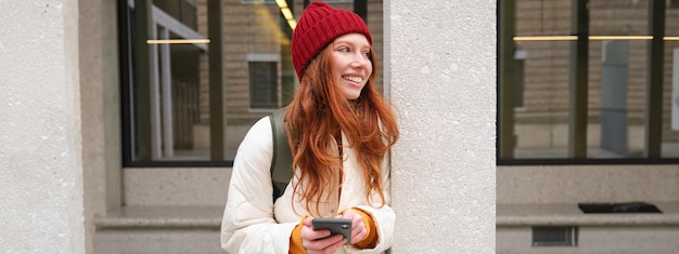 Beautiful redhead girl with smartphone standing on street holding mobile phone using taxi app