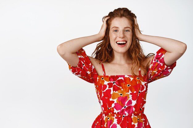 Beautiful redhead woman feeling carefree and happy holding hands on hair and smiling pleased standing in floral dress against white background