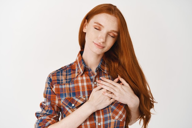 Beautiful redhead woman standing with closed eyes and hands on heart feeling touched dreaming about something standing over white background