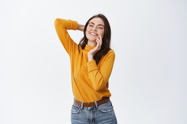 Beautiful relaxed woman hold hand behind head and touching natural beauty face, smiling and staring aside at copy space, standing carefree against white wall