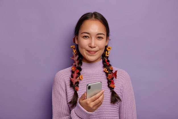 Beautiful season concept. Attractive Asian lady has natural beauty, dark hair combed in two plaits with fallen autumn leaves, enjoys autumnal coziness, uses modern smartphone during leisure time