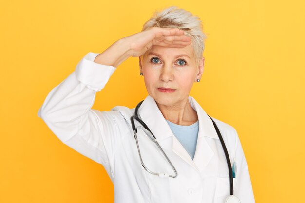 Beautiful senior woman doctor with dyed short hairstyle and blue eyes keeping hand on her forehead searching for something far away in distance.