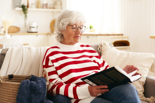 Beautiful senior woman with gray hair enjoying retirement, sitting on couch in living room, reading interesting novel. Elderly Caucasian female in round spectacles relaxing at home with book