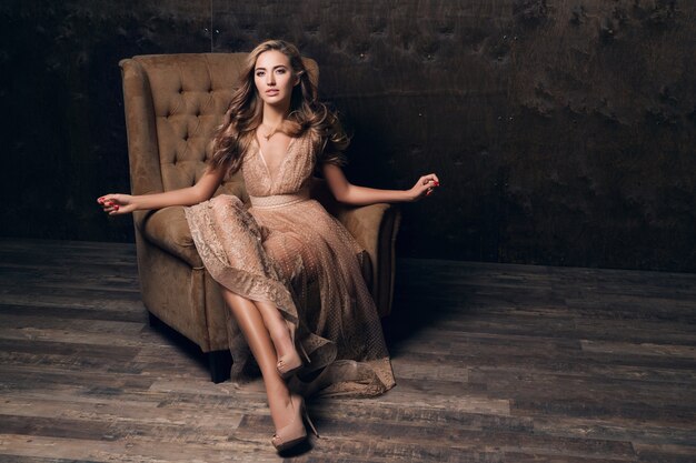 Beautiful sexy model woman in shiny lace evening dress posing sitting in chair of beige color