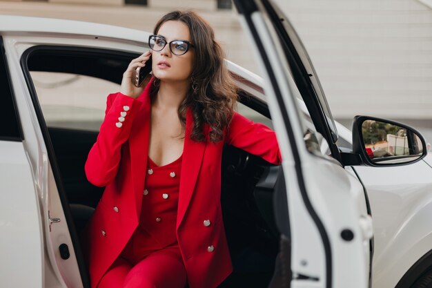 Beautiful sexy rich business woman in red suit sitting in white car, wearing glasses talking on phone, business lady style