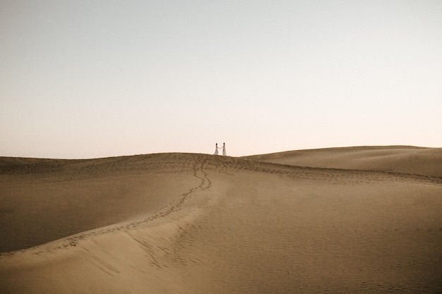 Beautiful shot of a desert hill with two females holding hands on the top in the distance