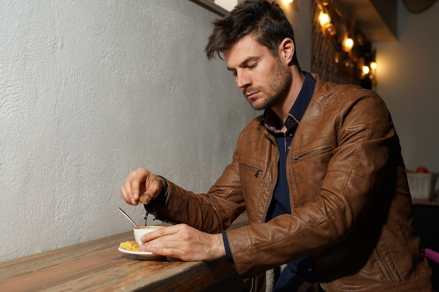 Free photo beautiful shot of an elegant male in a brown leather jacket adding sugar to his coffee