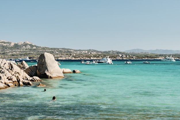 Beautiful shot of rocks in the water with boats and mountains in the distance under a blue sky