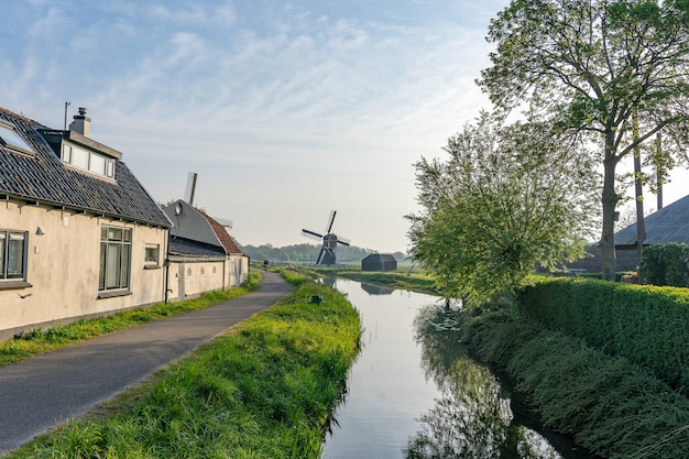 Beautiful shot of a water canal at the side of a narrow road with a windmill on a field