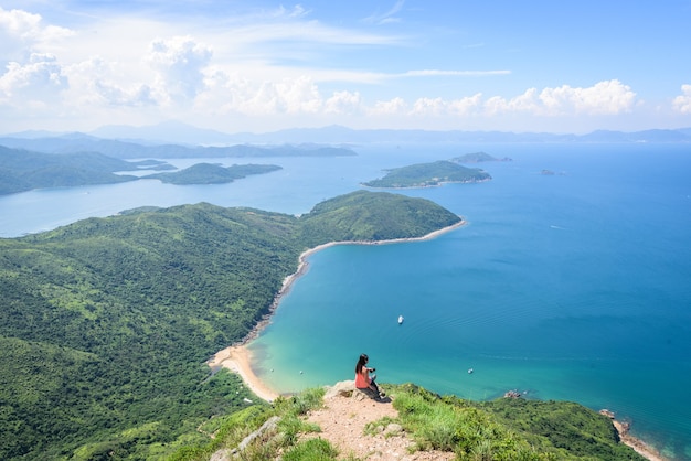 Free photo beautiful shot of a woman sitting on a cliff with a landscape of forested hills and a blue ocean