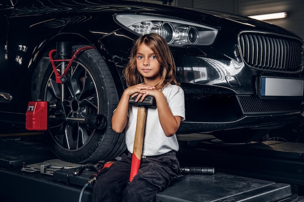 Beautiful small girl is sitting on the lifting platform with car and holding hammer.