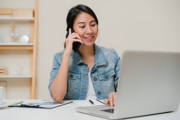 Beautiful smart business Asian woman in smart casual wear working on laptop and talking on phone while sitting on table in creative office. 