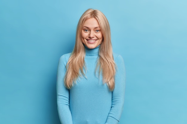 Beautiful smiling blonde woman expresses happiness being rejoiced at somebody has charming friendly face expression dressed in casual turtleneck 