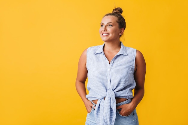 Beautiful smiling girl in shirt and jeans happily looking aside over yellow background isolated