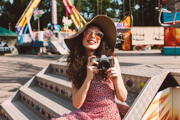 Beautiful smiling girl with dark curly hair in sunglasses and hat sitting with little camera in hand while happily spending time in amusement park