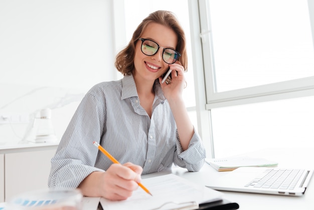 Beautiful smiling woman in glasses talking on mobile phone while working with documents at home