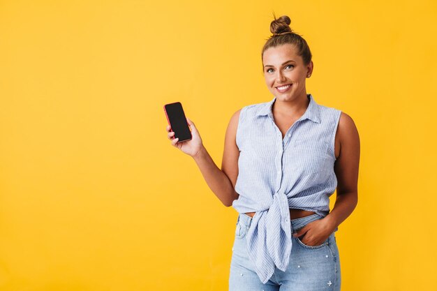 Beautiful smiling woman in shirt happily looking in camera while showing new cellphone over yellow background