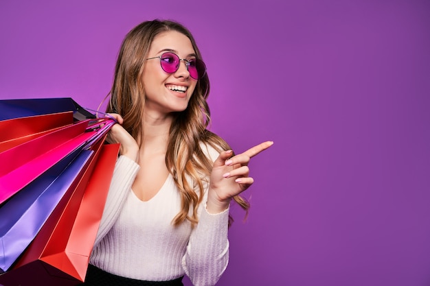 Free Photo beautiful smiling young blonde woman pointing in sunglasses holding shopping bags and credit card on a pink wall