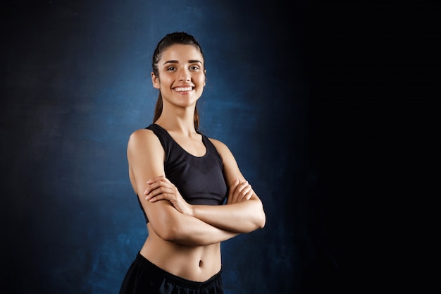 Beautiful sportive girl posing with crossed arms over dark wall.