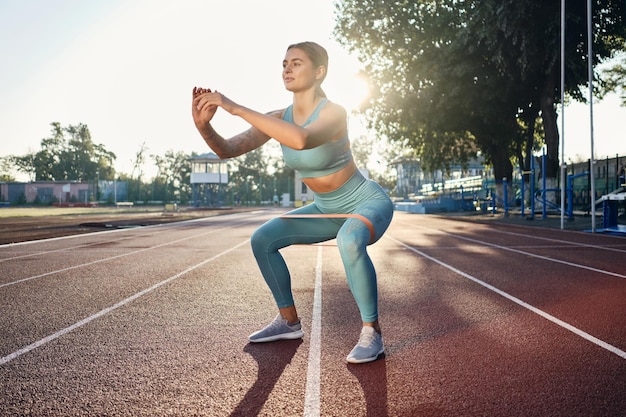 Beautiful sporty girl in stylish sportswear squatting with rubber band during workout on city stadium