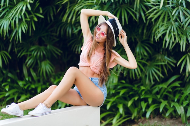 Beautiful stylish girl sitting on white fence in tropical park with stretched legs.