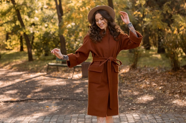 Free photo beautiful stylish smiling skinny woman with curly hair walking in park dressed in warm brown coat, autumn trendy fashion street style